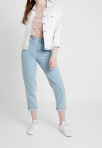 Biała kurtka dżinsowa, jasnoróżowy sweter w paski, jasnoniebieskie jeansy z podwiniętymi nogawkami i białe sneakersy. Neutralne tło.