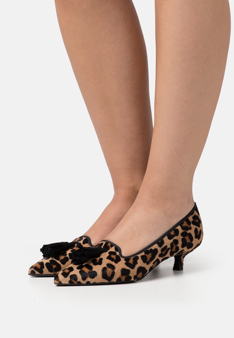 Chaussures à talons bas à bout pointu imprimé léopard avec des pompons noirs, portées pieds nus sur un fond uni.