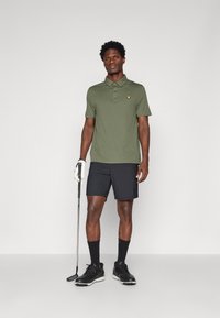 Camisa polo verde-escuro com um logo amarelo, combinada com calções pretos. O modelo segura um taco de golfe e usa sapatos pretos e meias até ao tornozelo.