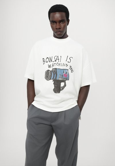 T-shirt bianco oversize con un design disegnato a mano di una macchina fotografica e la scritta "Bonsai ti sta osservando". Abbinato a pantaloni grigi.