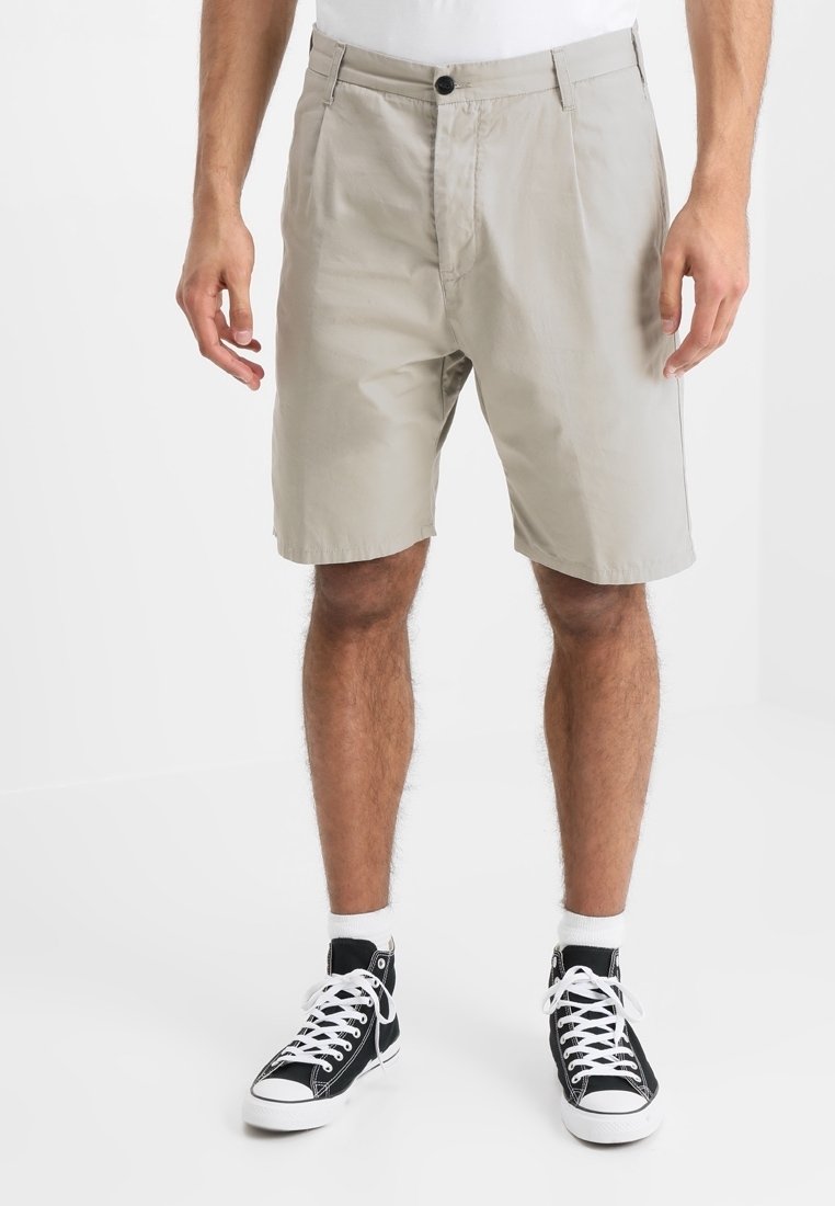 Shorts beiges en tissu mélangé coton, avec une finition plate à l'avant, des passants de ceinture et une fermeture à bouton. Portés avec des baskets noires.