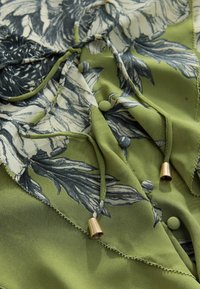 Bluza verde cu imprimeu floral, confecționată dintr-un material texturat, cu șnururi de ajustare cu accente aurii și nasturi decorativi. Model floral detaliat în tonuri delicate.