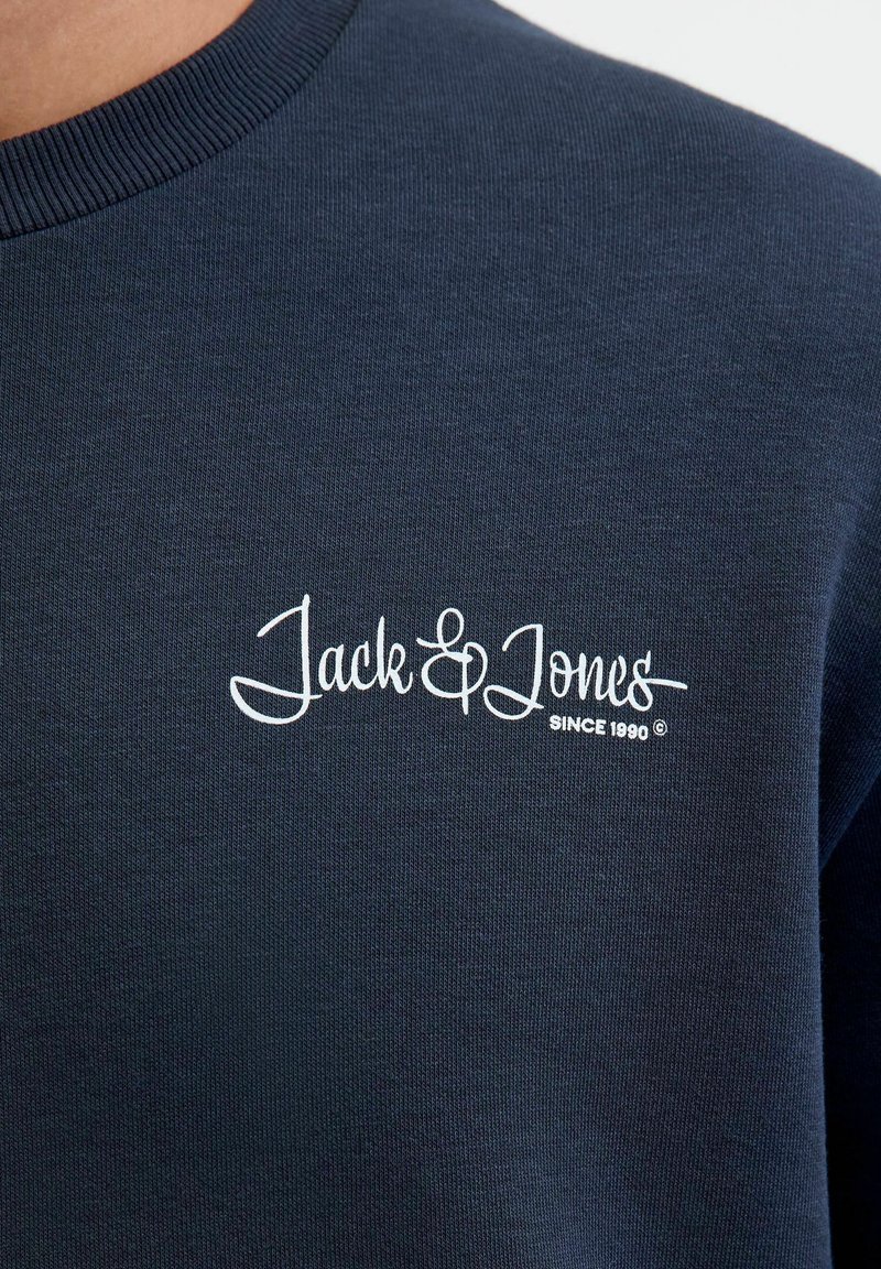 Marineblauwe sweatshirt met geribbelde halslijn; heeft witte geborduurde branding "Jack & Jones" en "SINCE 1990." Zachte textuur, casual ontwerp.