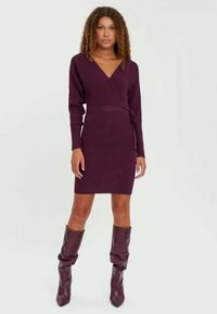 Knee-length, oprijetna obleka v temno vijolični barvi z V-izrezom in dolgimi rokavi, ki ima podrobnost z vezanimi pasom in rebrasto teksturo.