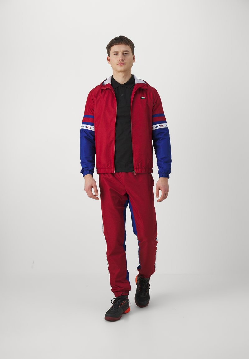 Rood en blauwe trainingspak met een ritsjack en elastische manchetten, gecombineerd met zwarte schoenen. Bevat logo-accenten en een sportief ontwerp.