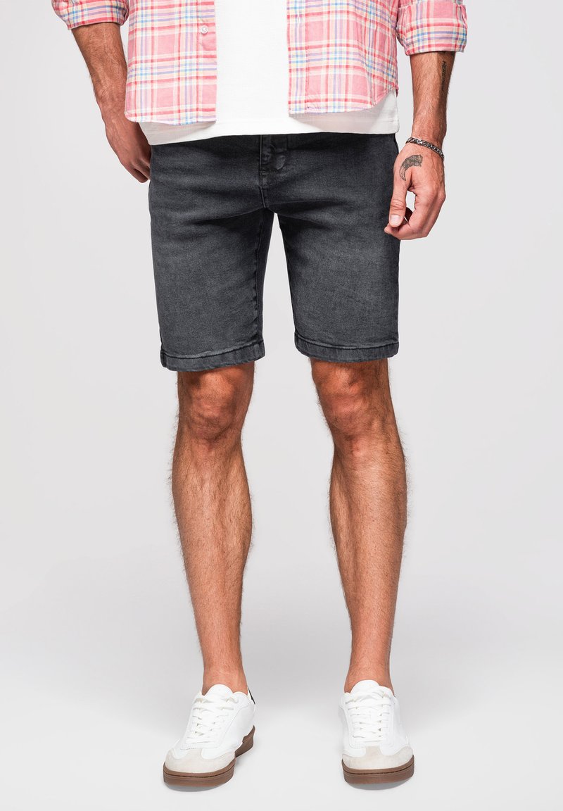 Mężczyzna stoi, ubrany w czarne jeansowe szorty, białe sneakersy, biały t-shirt i różową koszulę w kratę. Ręce swobodnie opuszczone wzdłuż ciała.