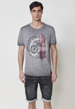 Camiseta gris de cuello redondo con un diseño desvanecido y un gráfico de motocicleta. Combinada con pantalones cortos negros con bolsillos de carga y dobladillos enrollados.