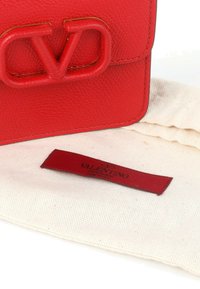 Bolso Valentino de piel roja con gran logo en relieve, reposando sobre una funda de tela crema con etiqueta roja Valentino Garavani.