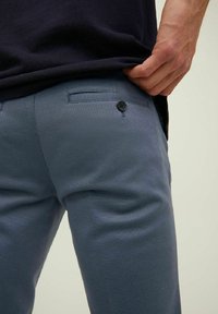 Hombre con pantalones azul oscuro que tiene un bolsillo trasero abotonado, levantando ligeramente una camisa azul marino con su mano derecha contra un fondo claro.