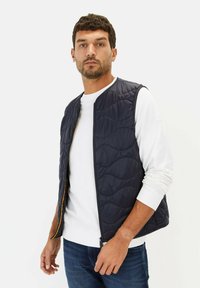 Marineblaue gesteppte Weste mit einem Reißverschluss und kontrastierendem orangefarbenem Futter, getragen über einem weißen Langarmshirt mit strukturierter Außenseite.