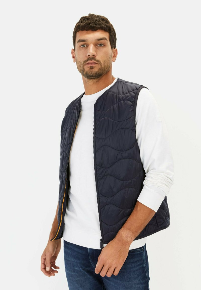 Marineblaue gesteppte Weste mit einem Reißverschluss und kontrastierendem orangefarbenem Futter, getragen über einem weißen Langarmshirt mit strukturierter Außenseite.