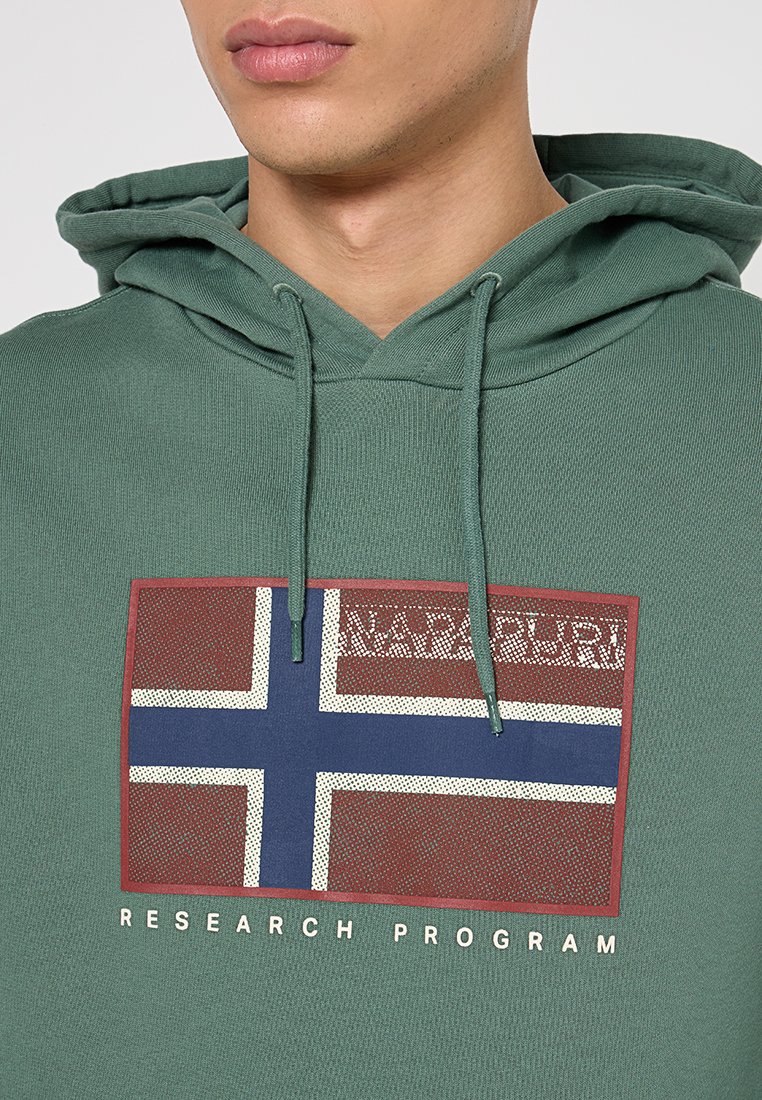 Mann trägt einen grünen Kapuzenpullover mit stilisiertem norwegischen Flaggenmotiv und dem Text "Napapijri Research Program" auf der Brust.