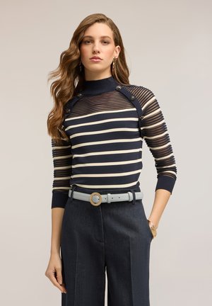 Frau mit welligem Haar, die ein transparentes Oberteil mit marineblauen und beigen Streifen und Knöpfen trägt, dazu einen grauen Gürtel, dunkle Hose und goldenen Schmuck, die Hände in den Taschen.