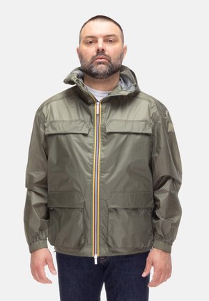 Uomo con capelli corti e barba che indossa una giacca con cappuccio verde oliva con molteplici tasche e una zip gialla prominente, in piedi davanti a uno sfondo bianco.