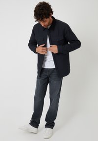 Marineblauwe knoopjack met een contrasterende kraag, twee voorzakken, gedragen over een wit shirt en gecombineerd met donkere jeans en witte sneakers.