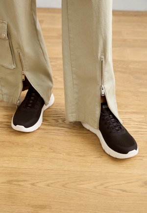 Mustad tekstuuriga mustad spordijalatsid valgete talladega on osaliselt kaetud heleda beeži värvi pükstega, millel on külgedel tõmblukud, mis ulatuvad alt üles.