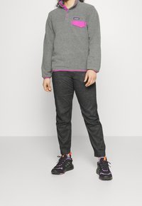 Harmaa fleece-pulloveri, jossa violetti tasku ja reunus, kauluksessa kolme nappia. Yhdistetty tummiin housuihin ja mustiin lenkkareihin, joissa värikkäitä yksityiskohtia.