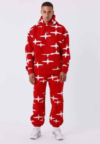 Sudadera roja y pantalones de chándal con patrones de estrellas blancas. El material es suave; la sudadera cuenta con un bolsillo frontal. El conjunto tiene un corte relajado y los pantalones son estrechos en los tobillos.