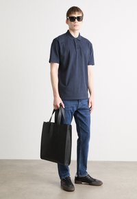 Bolsa tote preta de couro com duas alças, usada com uma polo azul marinho e jeans azuis. Modelo está posicionado contra um fundo neutro.