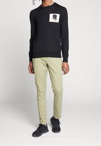 Black sweater with crew neck, featuring a white square patch displaying a purple rose and lettering. Paired with light green pants and black sneakers.