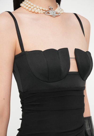 Sieviete, kas valkā melnu kleitu ar siksnām, strukturētiem kausiņiem un ruffles audumu, aksesuārus veido vairāklu pērlīšu kaklarotu ar sudraba zvaigznes kulonu.