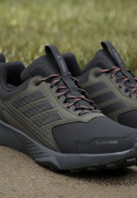 Schwarze und olivgrüne Trail-Schuhe mit strukturiertem Mesh-Obermaterial, leuchtend orangen Schnürsenkeln und einer robusten Gummisohle. Mit "CLIMAPROOF"-Logo.