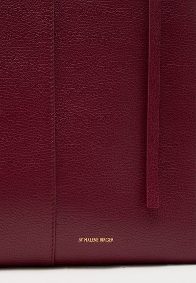 Τσάντα ώμου σε σκουρόχρωμο κόκκινο δέρμα με υφή επιφάνειας, με κεντρική ραφή και διακριτικό ενσωματωμένο λογότυπο της μάρκας σε χρυσό στο κάτω μέρος.