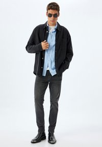 Chaqueta de mezclilla negra sobre una camisa de color azul claro, combinada con jeans oscuros y botas de tobillo negras. Presenta un bolsillo frontal y gafas de sol.