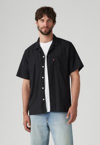 Zwart shirt met korte mouwen en een subtiele textuur, witte knopen en een zakje op de linkerborst, gedragen over een wit T-shirt, gecombineerd met lichtblauwe spijkerbroek.