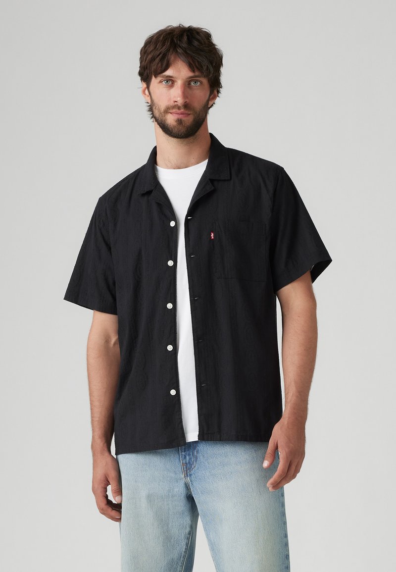 Camisa de manga corta negra con textura sutil, botones blancos y un bolsillo en el pecho izquierdo, usada sobre una camiseta blanca, combinada con jeans azul claro.