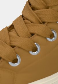 Sneaker marrone con tomaia testurizzata, lacci intrecciati, occhielli metallici e suola in gomma bianca. Vista ravvicinata che evidenzia i dettagli dei lacci.