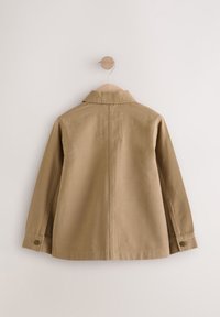 Veste en coton beige avec un empiècement au dos, manches longues et poignets boutonnés. Dotée d'une poche horizontale sur le haut du dos. Texture lisse.