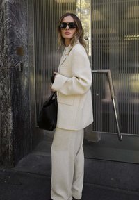 Blazer texturé beige avec un pantalon large assorti, accompagné de lunettes de soleil noires et d'un sac à main noir, sur fond moderne en verre et métal.