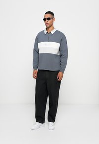 Grå zip-up sweatshirt med en hvid stribe og logo-detalje, parret med løse sorte bukser og hvide sneakers. Model iført mørke solbriller.