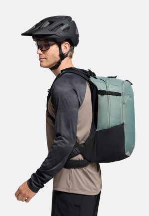 Zaino con finitura in tessuto verde, sezione inferiore nera, multiple compartimenti, spallacci regolabili e una persona che indossa un casco nero e occhiali da sole.