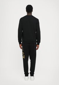 Svart hettegenser og joggebukser-kombinasjon. Hettegenseren har rund hals; joggebuksene har gullbokstaver. Begge er tettsittende og laget av mykt materiale.