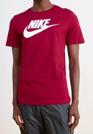 Mann trägt ein rotes T-Shirt mit kurzen Ärmeln, großem weißem Nike-Logo und schwarze Shorts vor einfachem hellem Hintergrund.