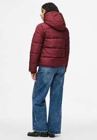 Burgundyröd puffjacka med huva, vadderad design och åtsittande silhuett. Matcher med hög midja, vida blå jeans och svarta stövlar.