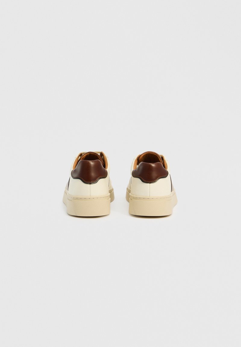 Sneaker mit einem cremefarbenen synthetischen Obermaterial, braunen Lederakzenten an der Ferse, beigefarbenen Gummisohlen und glatter Textur. Klassisches Schnürdesign.