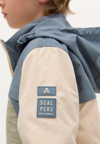 Veste bloc de couleurs avec un design bleu, beige et vert. Comprend une capuche et des patches en caoutchouc sur la manche. Texture lisse.