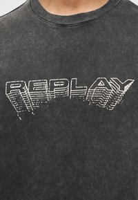 Κοντινό πλάνο ενός σκουρογκρι T-shirt με τη λέξη "REPLAY" σε λευκά, περιγραμματικά 3D γράμματα πάνω στο στήθος.