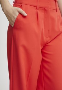 Red tailored high-waisted trousers with a smooth texture, featuring side pockets and neat pleats. Visible stitching and clean lines.