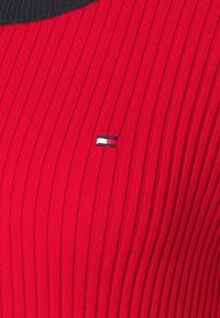 Pull en maille côtelée rouge avec un col noir et un petit logo brodé en rouge, blanc et bleu sur le côté gauche.