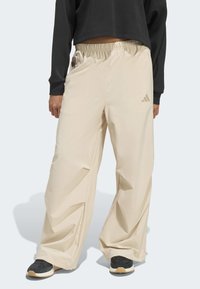 Béžové volné kalhoty s elastickým pasem, s bočními kapsami a nenápadným logem Adidas, doplněné černými teniskami.