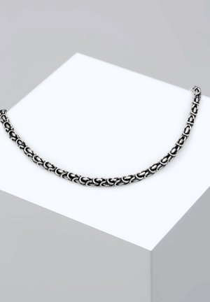 Zilveren ketting met schakels van onderling verbonden ovale ringen met een gepolijste afwerking, tentoon gesteld op een witte kubus als standaard.
