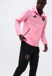 Άνδρας που φοράει ροζ μπουφάν με φερμουάρ και μαύρο παντελόνι με λογότυπα της ποδοσφαιρικής ομάδας του Μαϊάμι, ρυθμίζοντας την τσέπη του μπουφάν, σε απλό λευκό φόντο.