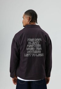 Donker geruit overshirt met geborduurde tekst, voorzien van een kraag, lange mouwen en een losse pasvorm. De achtergrond is egaal grijs.