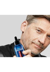 Bouteille compte-gouttes en verre bleu étiquetée "L'Oréal Men Expert Power Age", tenue par un homme. Fond gris clair ; le visage de l'homme affiche des lignes d'application de soins de la peau en points.