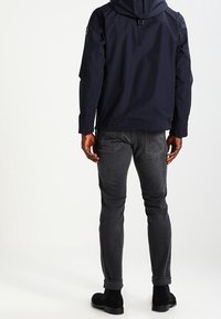 Marineblå vandtæt jakke med hætte, med lynlås-lukning og diskrete skulderdetaljer, parret med slim-fit mørkegrå jeans og sorte fodtøj.