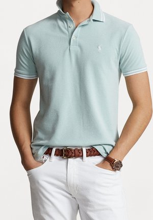 Homme portant un polo bleu clair, un pantalon blanc, une ceinture tressée marron et une montre à bracelet en cuir marron, les mains dans les poches.